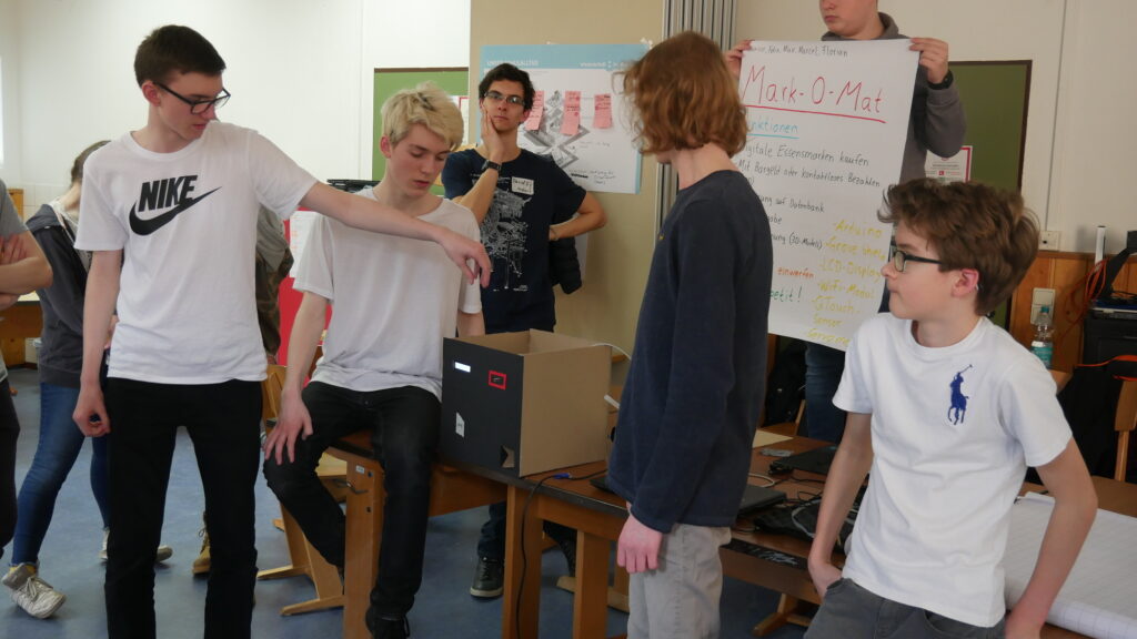Eine Gruppe Schüler steht um einen selbstgebauten Pappkasten herum und zeigt die Funktionen des Prototyps.