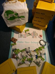 Auf dem Bild ist ein Plakat von Make Your School mit einzelnen Bestandteilen des Material-Kits zu sehen. Neben den gepackten Boxen ist ein Stoffelbeutel vom LJBW zu erkennen.