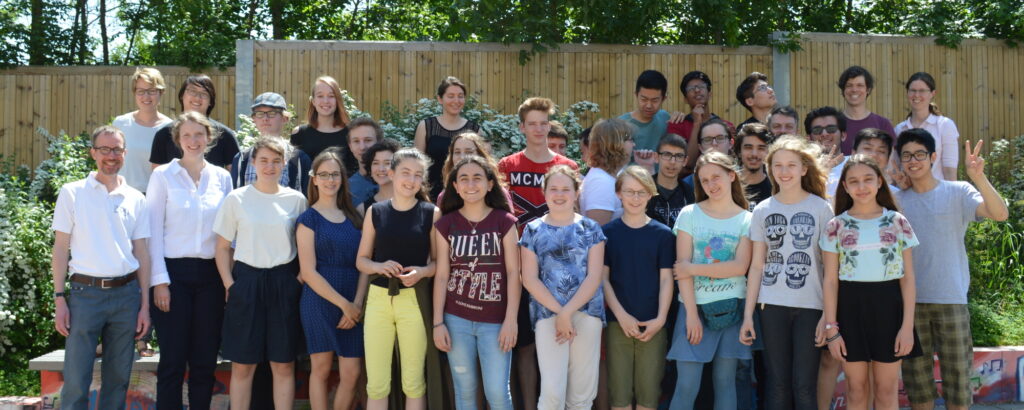 Etwa 30 Jugendliche, vier junge Erwachsene und drei Lehrkräfte posieren lächelnd für ein Gruppenfoto. Sie stehen im Pausenhof der Schule.