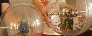 Zwei Jugendliche halten jeweils einen basketballgroßen Plastikball in die Kamera. Im Inneren der Bälle sind technische Bauteile an einer Stange fixiert, die quer durch den jeweiligen Ball läuft.