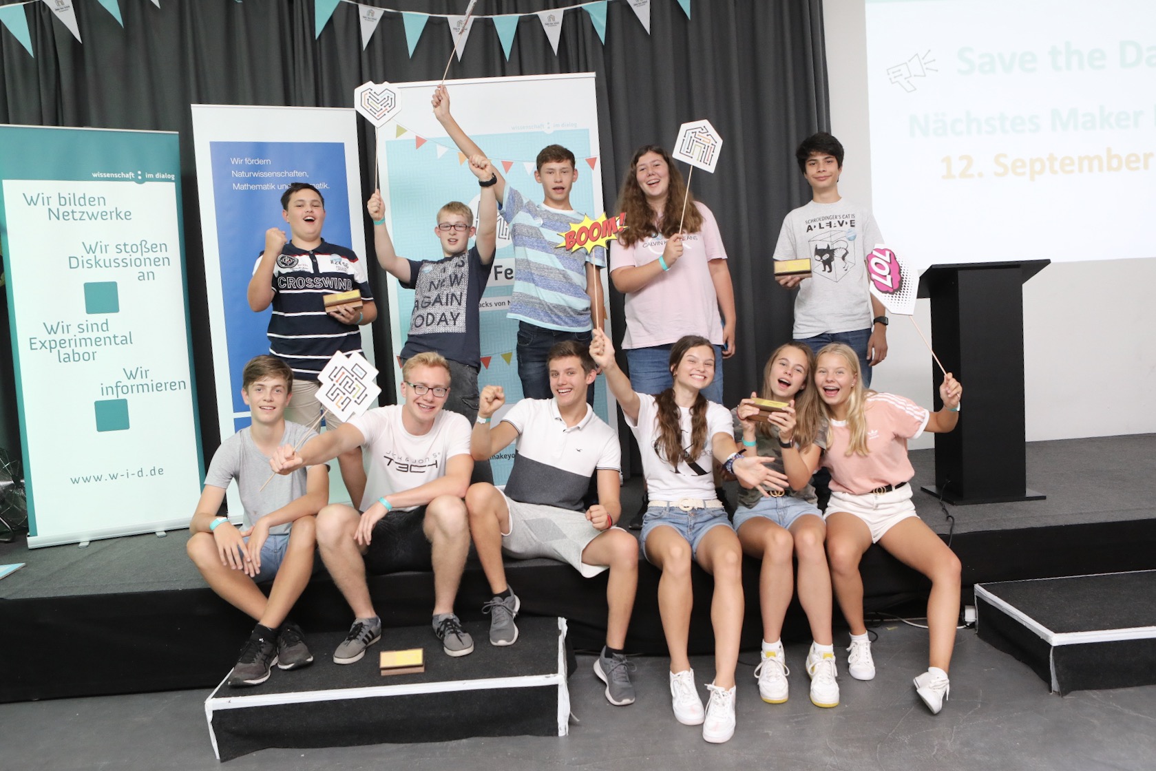 Jugendliche strahlen und lachen auf einer Bühne. Sie halten Schilder mit den Logos von Make Your School in die Kamera.