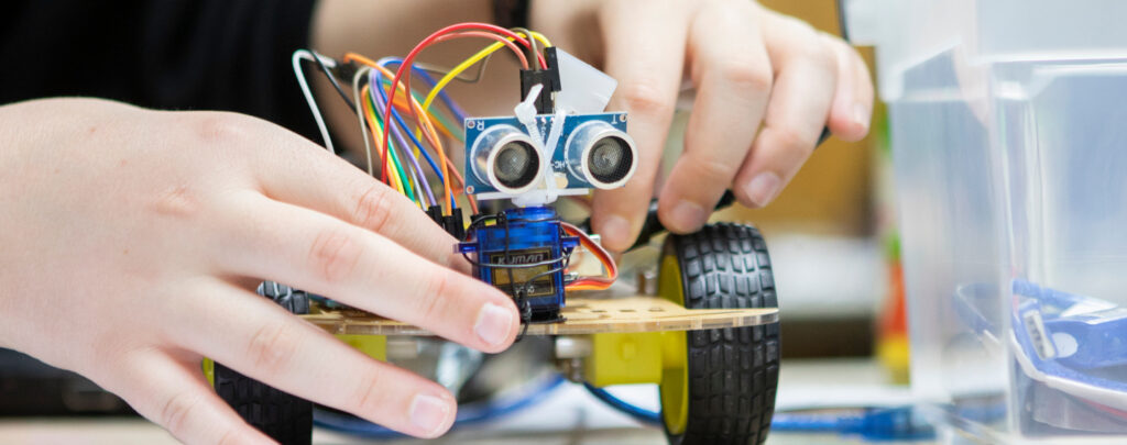 Zwei Hände basteln an einem kleinen gelben Roboterauto. Zwei Sensoren an seiner Vorderseite sehen aus wie Augen. Aus seinem Kopf kommen Drähte.