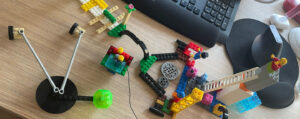 Das Bild zeigt einen Schreibtisch von oben. Darauf ist mit Legosteinen etwas durcheinander eine kleine Landschaft gebaut. Zu erkennen sind eine Legofigur auf einem Sprungturm, eine Art Zirkusdirektor und – mit etwas Fantasie – eine Wiese mit einer Blume.