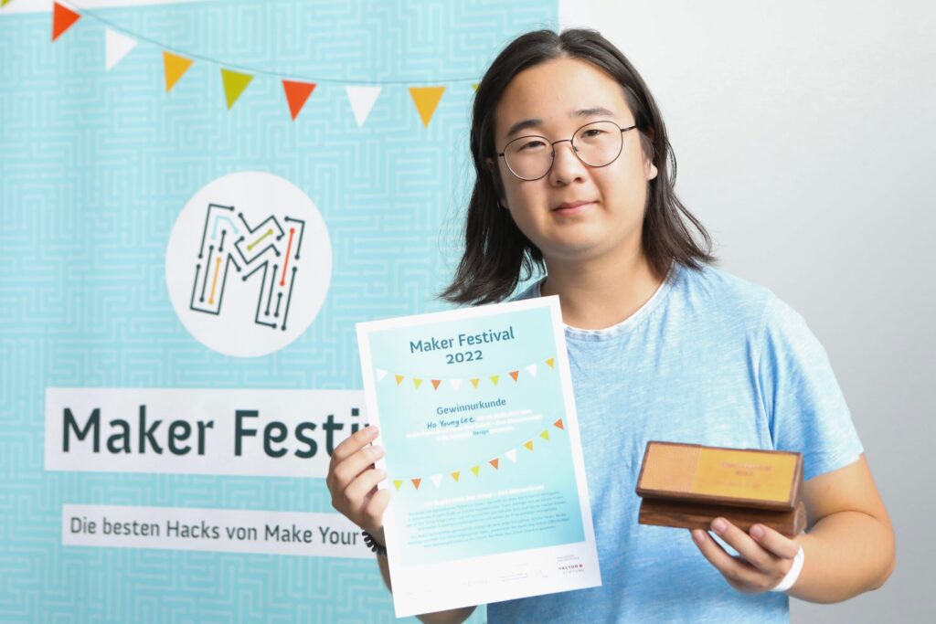Ein Jugendlicher steht vor einem blauen Maker-Festival-Plakat. In der linken Hand hält er einen kleinen, flachen Holzpokal in Form eines rechteckigen Quaders. In der rechten Hand hält er seine Maker Festival 2023 Urkunde.
