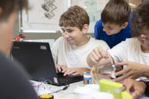 Mehrere Schüler sitzen um einen Computer herum. Einer der Schüler hält Kabel in der Hand.