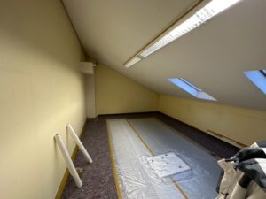 Ein leerer Raum mit schräger Decke. Die Wände sind hellgelb. Der Boden ist mit Folie und Malerflies ausgelegt.