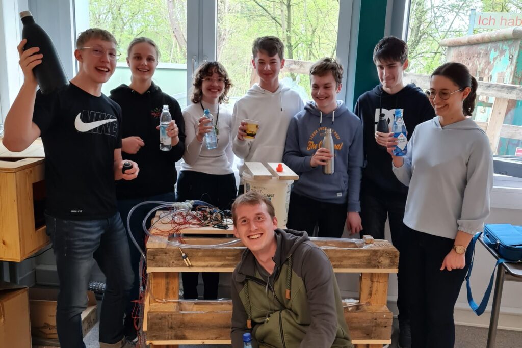 Sieben Jugendliche stehen hinter einer Holzpalette und lachen in die Kamera. Vor der Bank kniet ein junger Mann und lächelt ebenfalls in die Kamera.