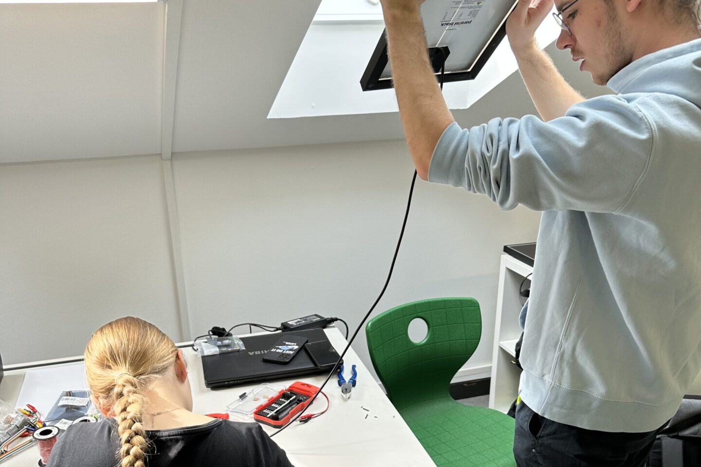 Ein Mädchen sitzt an einem Tisch und senkt ihren Kopf konzentriert nach unten. Ein Junge steht neben ihr am geöffneten Dachfenster und hält das Photovoltaikmodul Richtung Himmel.