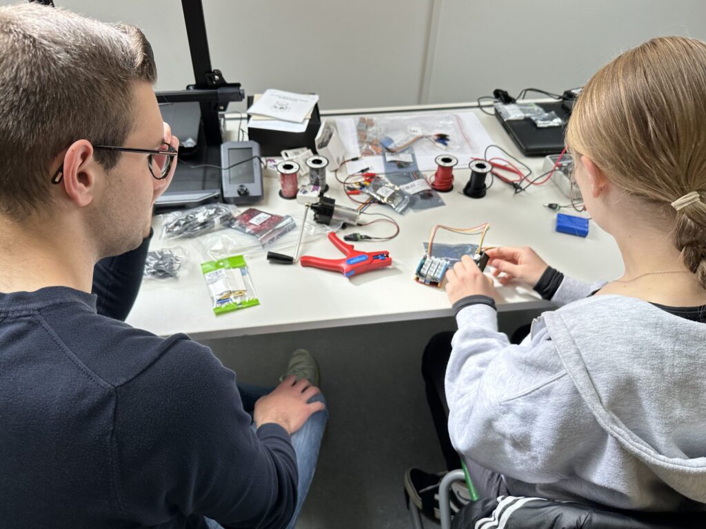 Ein Lehrer sitzt mit einer Schülerin am Tisch, vor ihnen viele Kabel, Werkzeuge und technische Bauteile. die Schülerin steckt Kabel zusammen, der Lehrer schaut ihr dabei zu.