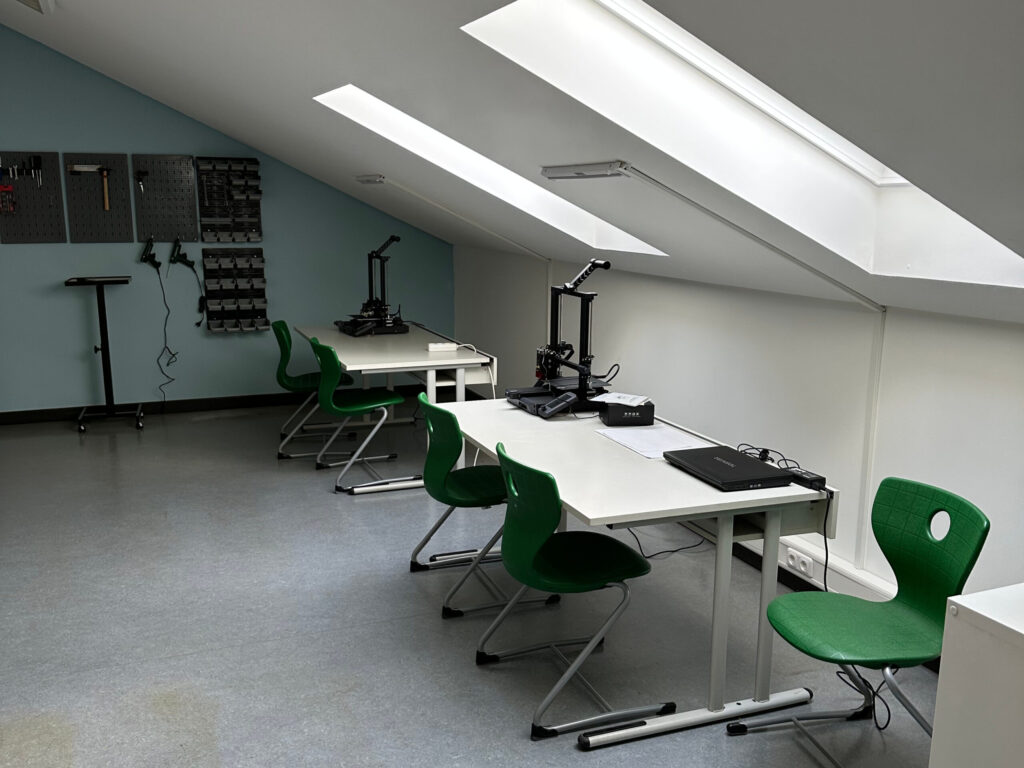 In einem Raum stehen Tische und Stühle. Darauf stehen technische Geräte. An einer Wand im Hintergrund hängt Werkzeug.