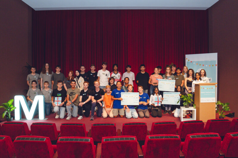 Mehrere dutzend Jugendliche stehen auf der Bühne für ein Gruppenfoto. Einige halten ein A2-großes Plakat. Bühne ist dunkelrot, vor der Bühne sind dunkelrote Kinostühle