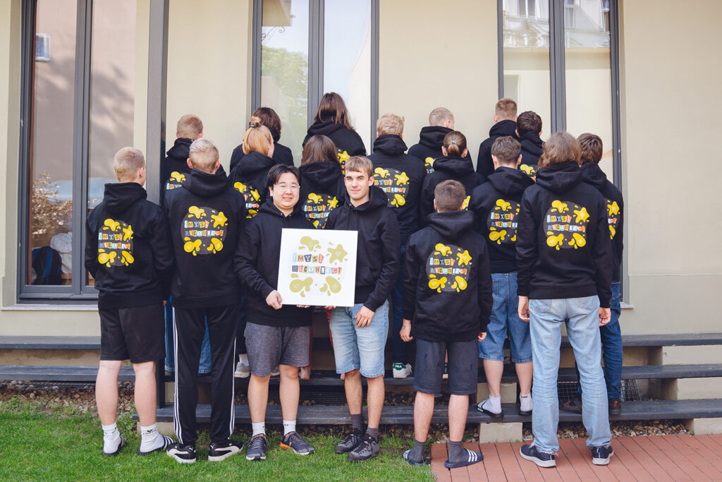 Gruppenbild von Jugendlichen. Fast alle stehen mit dem Rücken zur Kamera und tragen einen schwarzen Pullover mit dem neuen bunten mys reconnect Logo darauf. Zwei Jugendliche schauen in die Kamera und halten ein Schild, auf dem ebenfalls das neue Logo zu sehen ist.