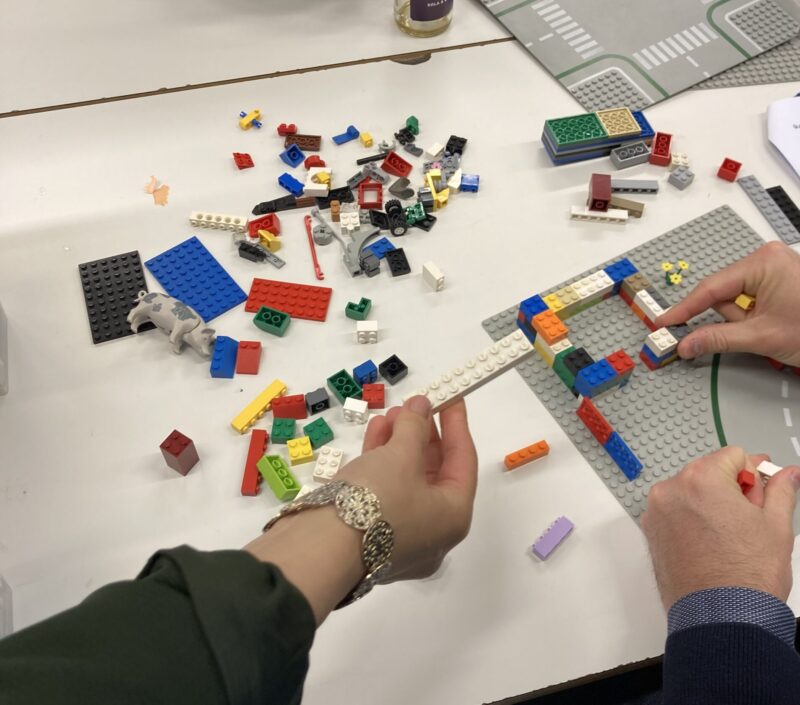Zwei Hände von 2 Personen vor einer weißen Tischplatte voller Legosteine, die gemeinsam etwas bauen