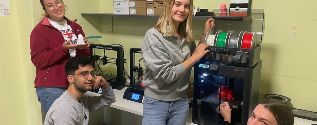 Vier Jugendliche, zwei weiblich gelesene und zwei männlich gelesene Personen stehen vor einem Tisch. Auf dem Tisch sind viele technische Geräte zu sehen.