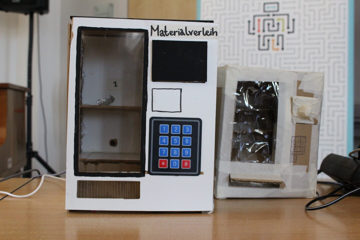 Ein selbstgebauter Kasten aus Pappe in der Größe eines großen Schuhkartons hat vorne ein kleines Desplay, ein Eingabefeld mit Zahlen, ein Fach, aus dem Materialentnommen werden kann. Darauf steht "Materialverleih".