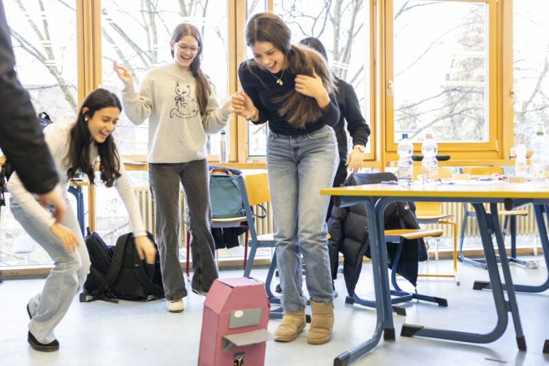 Vier Schülerinnen lachen und freuen sich in einem Klassenzimmer. Sie blicken auf den Boden, aus dem ein selbstgebauter pinkfarbener Roboter fährt.