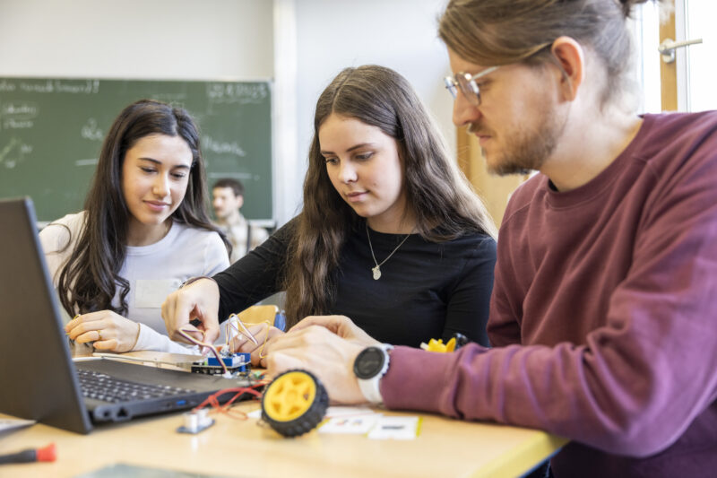 Zwei junge Frauen sitzen für einem Laptop und stecken Kabel an ein technisches Bauteil. Recht neben ihnen sitzt ein junger Mann und schaut auf die Steckverbindung.