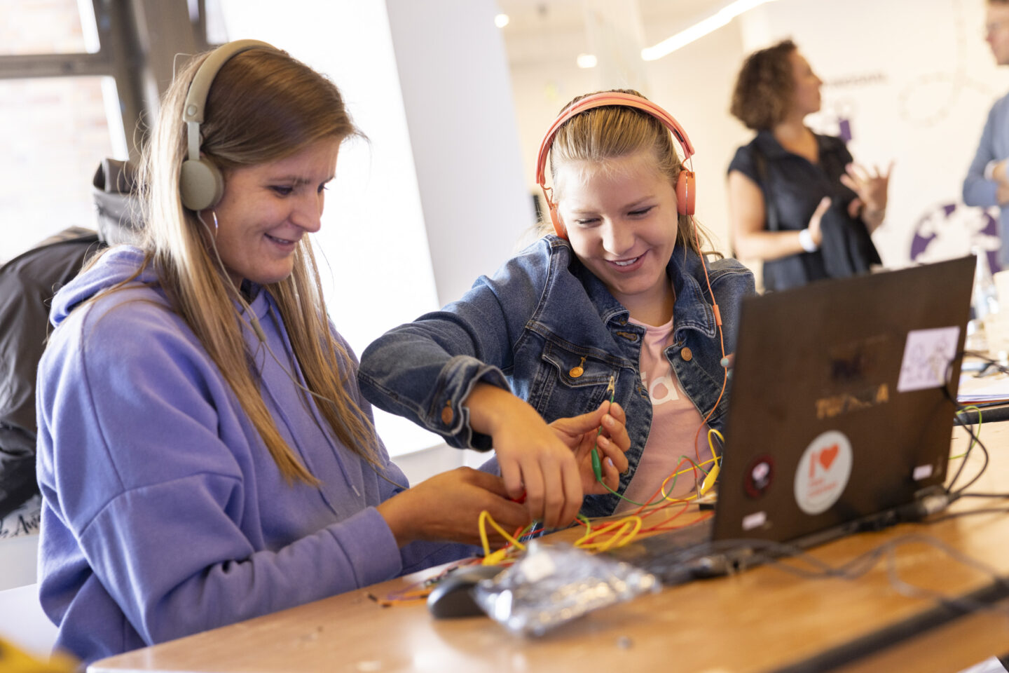 Zwei weiblich gelesene Jugendliche sitzen an einem Tisch und tüfteln mit elektronischen Bauteilen. Auf dem Tisch sind Kabel und ein Laptop.