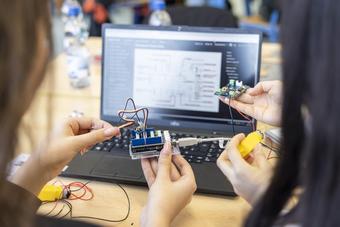 Auf einem Laptop ist eine technische Zeichnung zu sehen. Davor haben Hände einen Mikrocontroller hoch, aus dem einige Kabel zu einem kleinen Motor laufen.