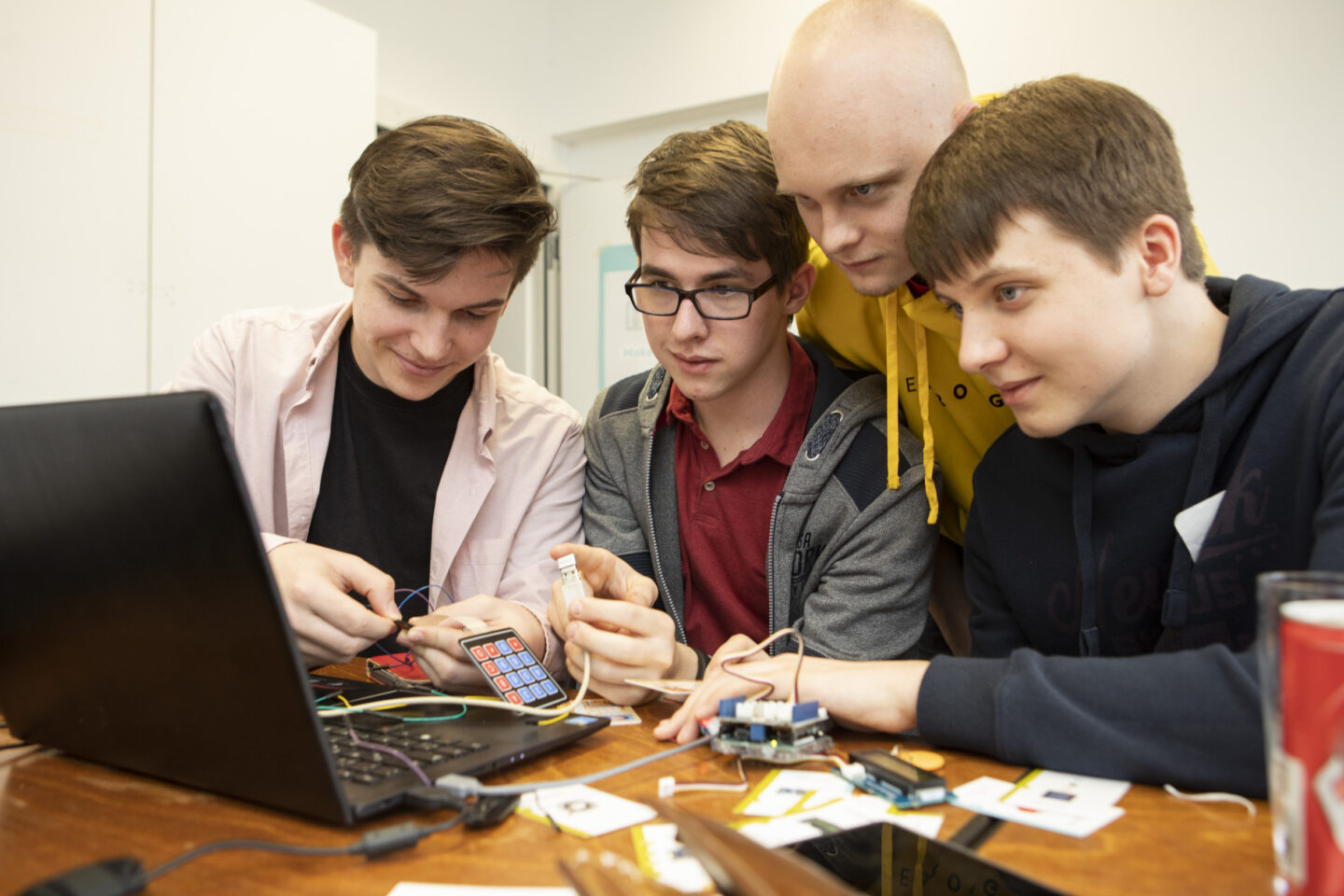 Vier Schüler sitzen vor einem Laptop. Davor liegen Kabel und zusammengesteckte Technik. Sie schauen konzentriert auf den Bildschirm.