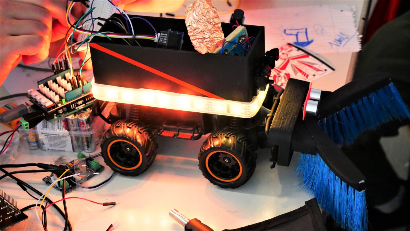 Auf einem Tisch steht ein schwarzer selbstgebauter Roboter. Er hat vier Räder, rundherum einen leuchtende LED-Streifen und zwei Bürsten vorne dran.