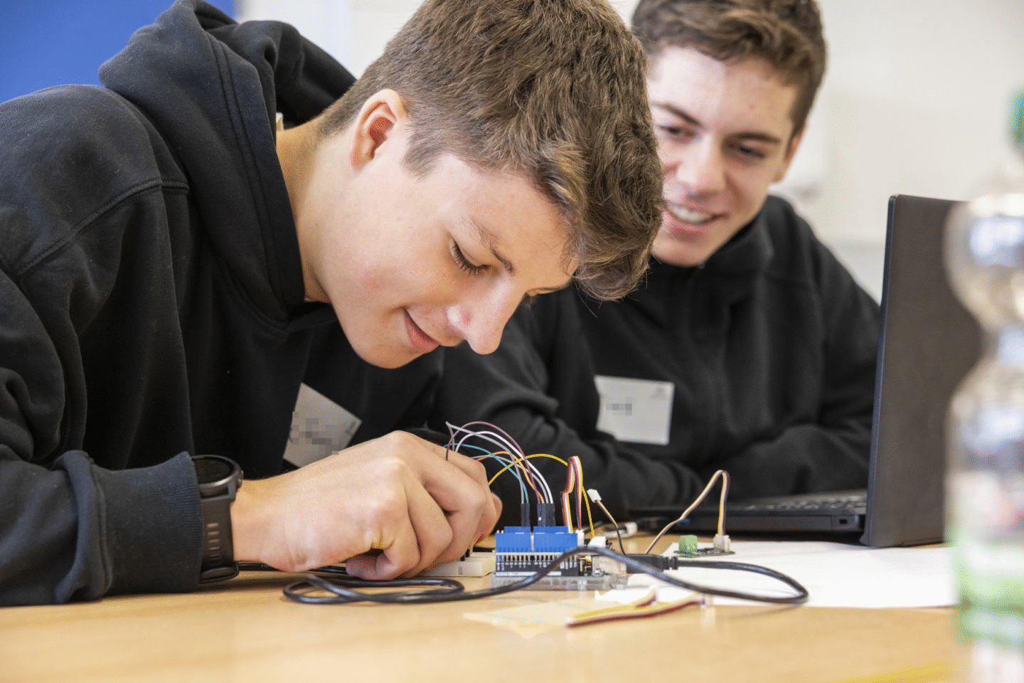Ein männlich gelesener Jugendlicher arbeitet am Tisch an technischen Bauteilen. Rechts daneben sitzt ein weiterer Jugendlicher der lächelt und zuschaut.