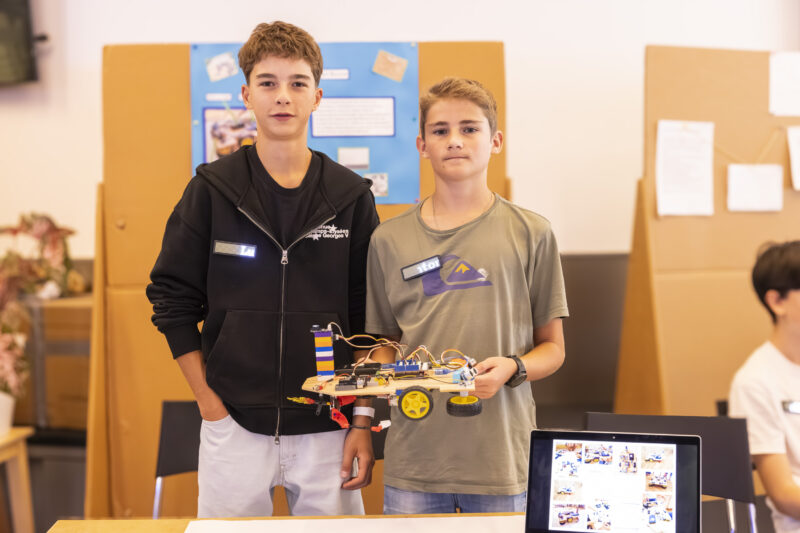 Zwei Jugendliche (Jungen) stehen vor dem Tisch und schauen in die Kamera, in der Hand halten sie ihren Prototypen.