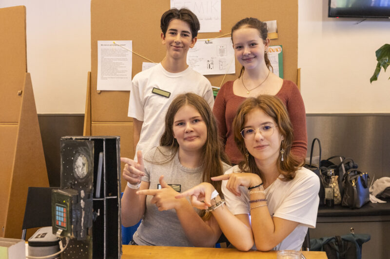 Vier Jugendliche (3 weiblich gelesen, 1 männlich) sitzen vor einem Tisch und schauen in die Kamera. Zwei zeigen auf einen Prototypen, der auf dem Tisch liegt.