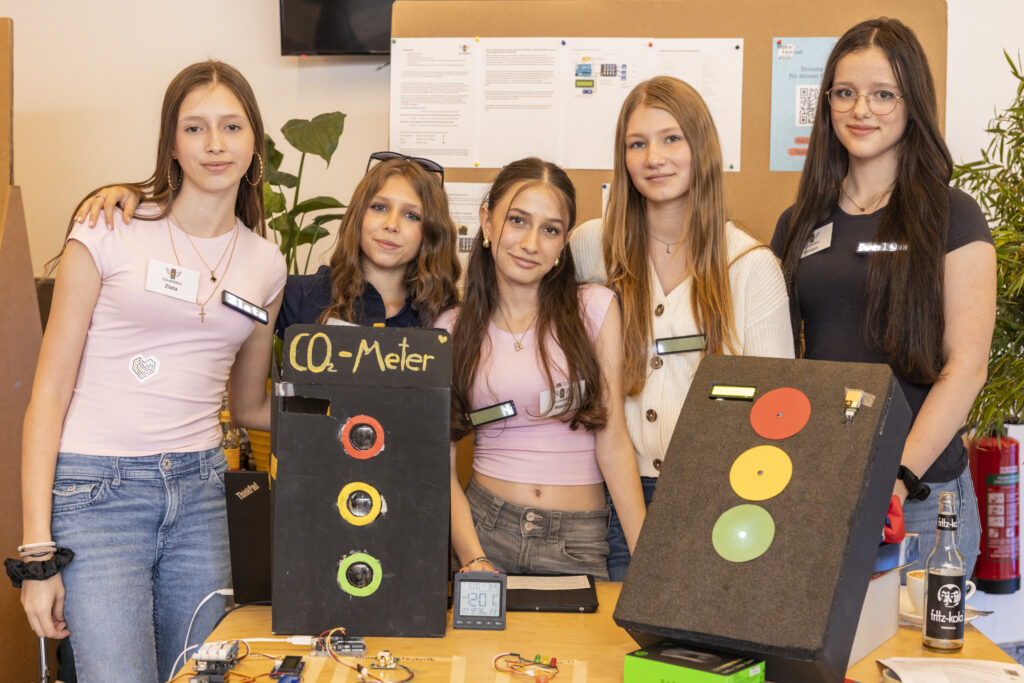 Fünf Jugendliche (Mädchen) stehen vor einem Tisch und schauen in die Kamera. Vor ihnen auf dem Tisch stehen zwei Prototypen.