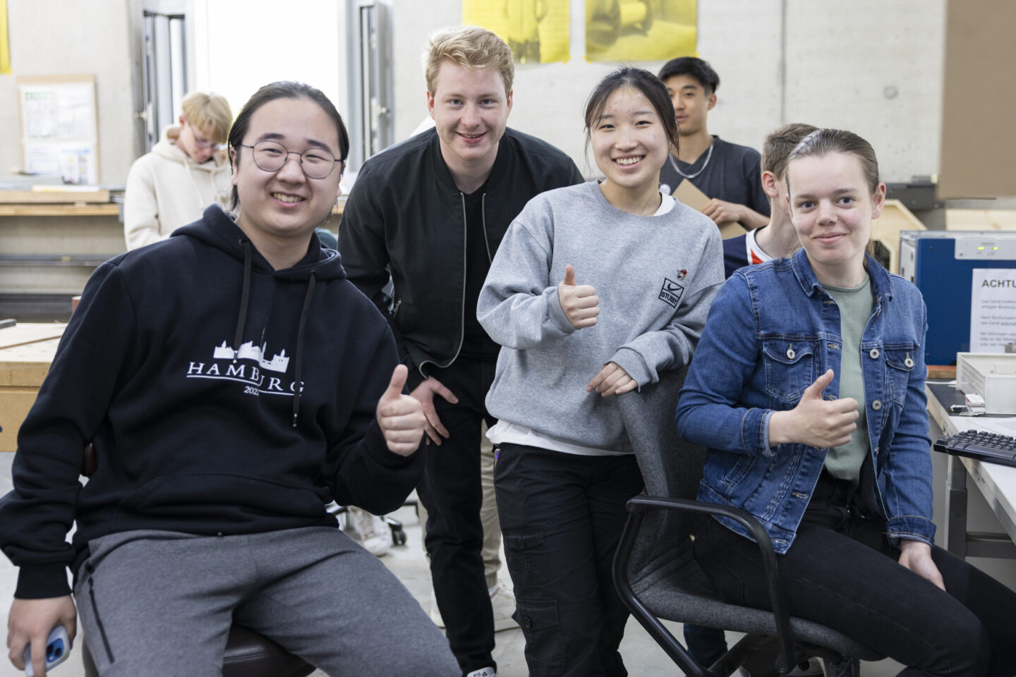 Zwei weiblich und zwei männlich gelesene Jugendliche sitzen und stehen nebeneinander, lächeln in die Kamera und zeigen Daumen nach oben.