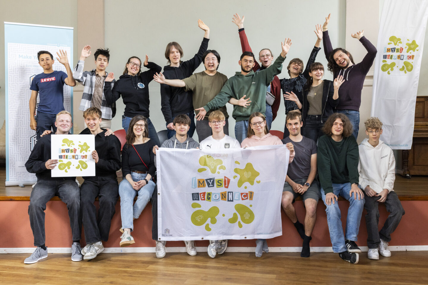 Eine große Gruppe an Menschen, hauptsächlich Jugendlicher, schauen in die Kamera und lächeln. Die obere Reihe hebt ihre Arme hoch. Die untere Reihe sitzt, ein paar Jugendliche halten ein Schild und ein Banner mit dem [mys]reconnect-Logo hoch.