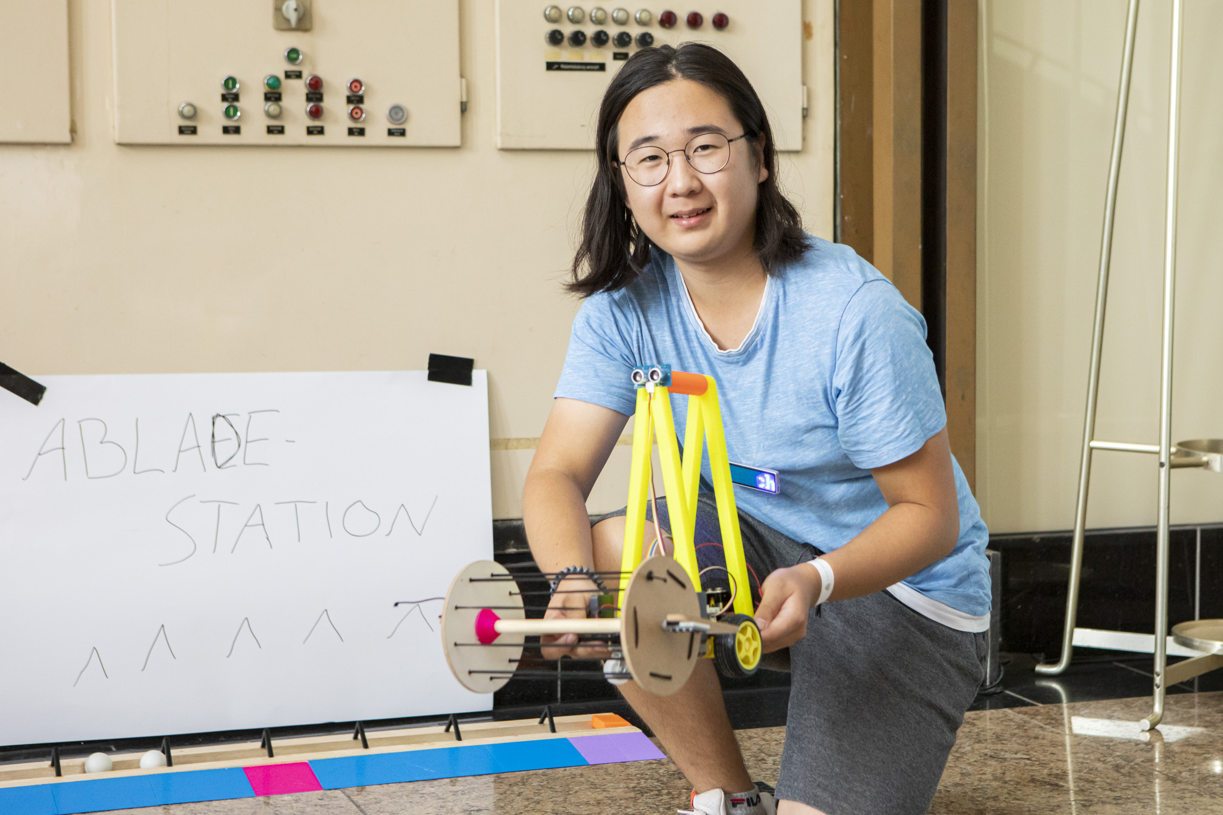 Ein männlich gelesener Jugendlicher steht in der Mitte des Bilds und hält seinen Roboter hoch und lächelt in die Kamera.