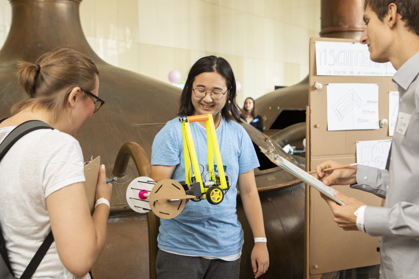 Ein männlich gelesener Jugendlicher steht in der Mitte des Bilds und hält seinen Roboter hoch. Jurymitglieder stehen links und rechts von ihm und begutachten den Roboter.