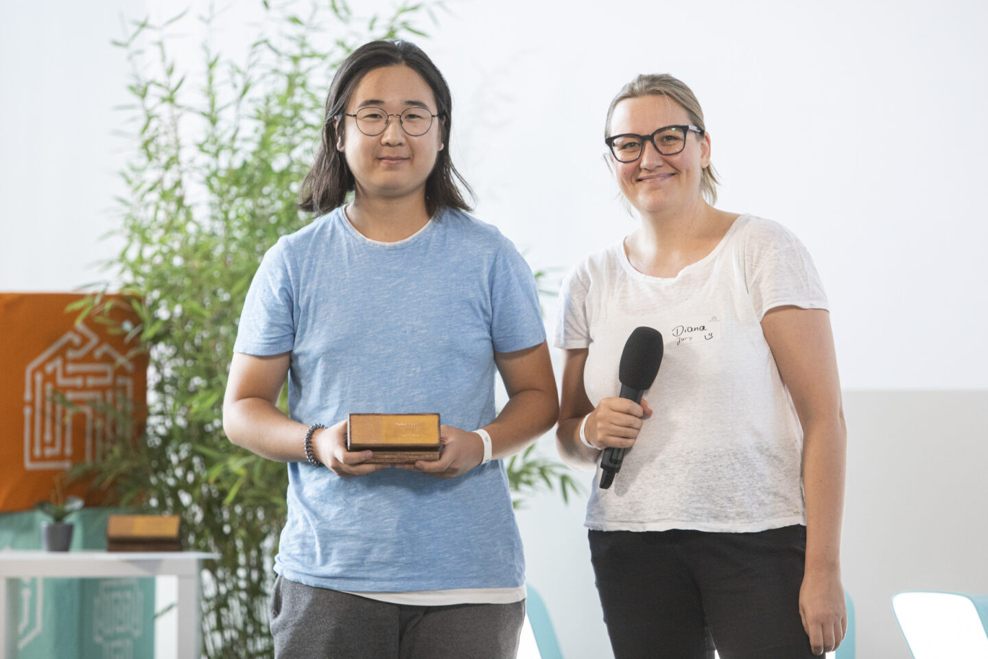 Ein männlich gelesener Jugendlicher hält einen Make-Your-School-Award in die Kamera. Daneben steht ein weiblich gelesenes Jurymitglied mit Mikrofon in der Hand. Beide lächeln in die Kamera.