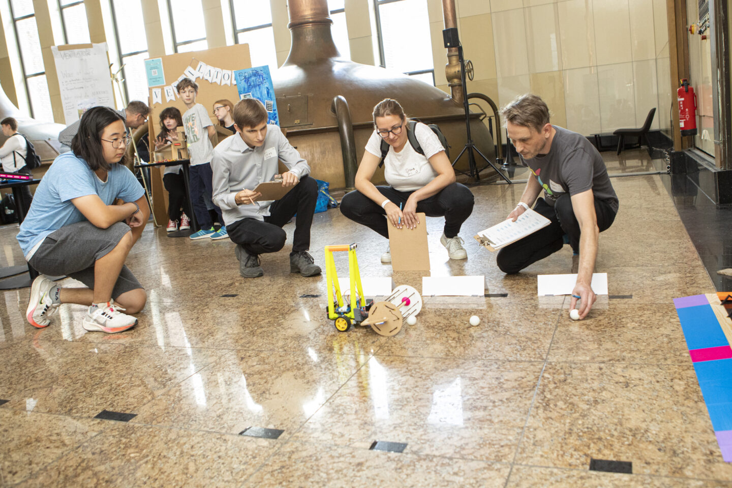 Ein männlich gelesener Jugendlicher und drei Jurymitglieder knien auf dem Boden und probieren einen Roboter aus, der auf dem Boden entlangfährt.
