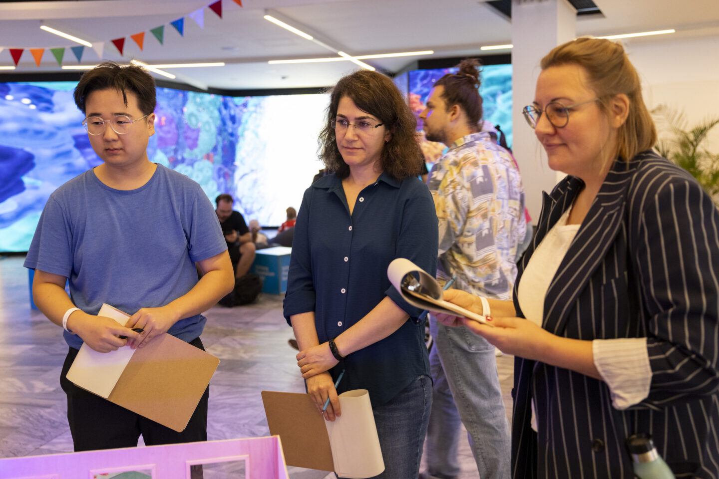 Drei Jury-Mitglieder stehen nebeneinander und halten jeweils ein Klemmbrett. Sie schauen in die gleiche Richtung.