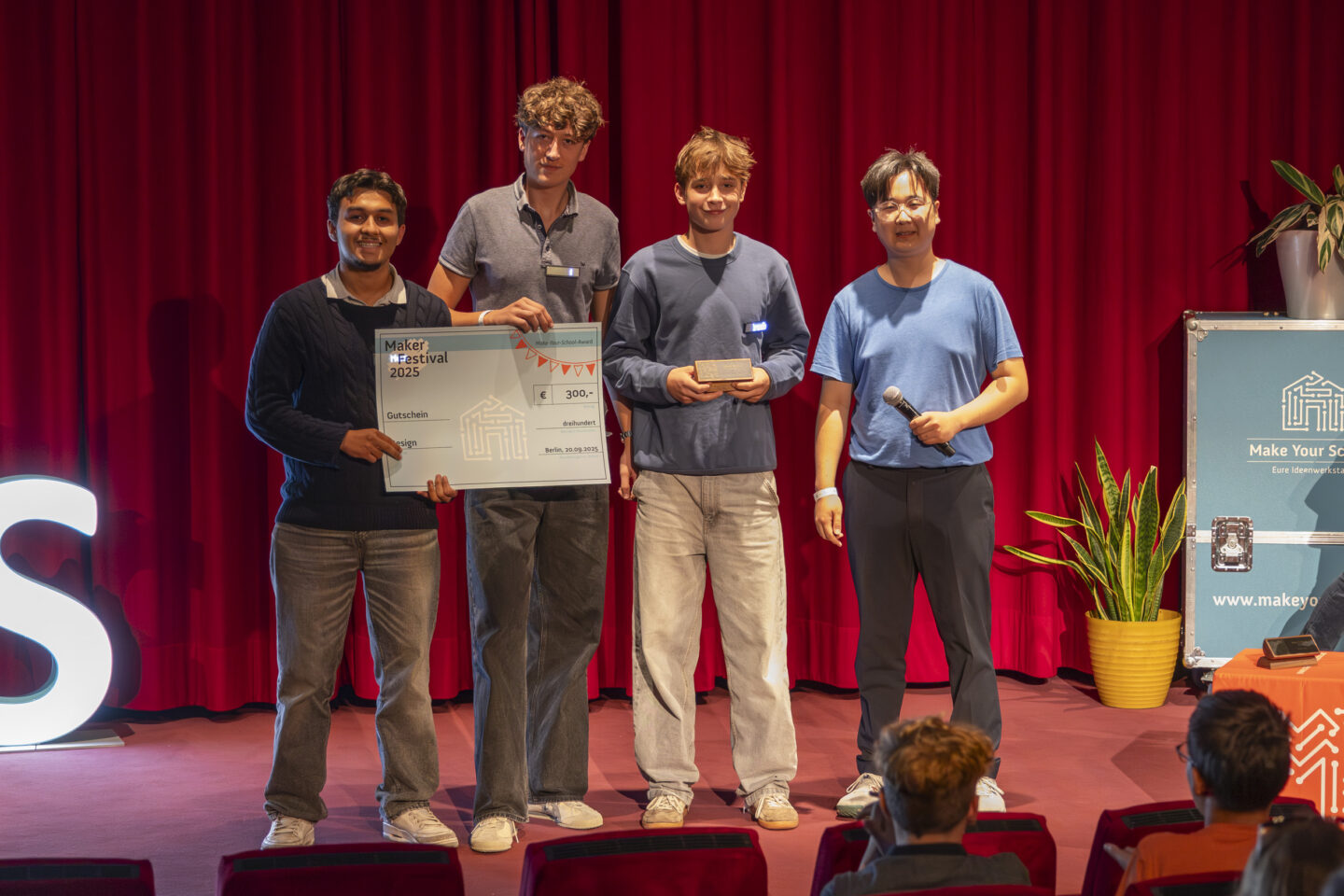 Drei Jugendliche und ein Jurymitglied stehen vor einem dunkelroten Vorhang, lächeln in die Kamera und halten einen großen Gutschein und den Make-Your-School-Award hoch.