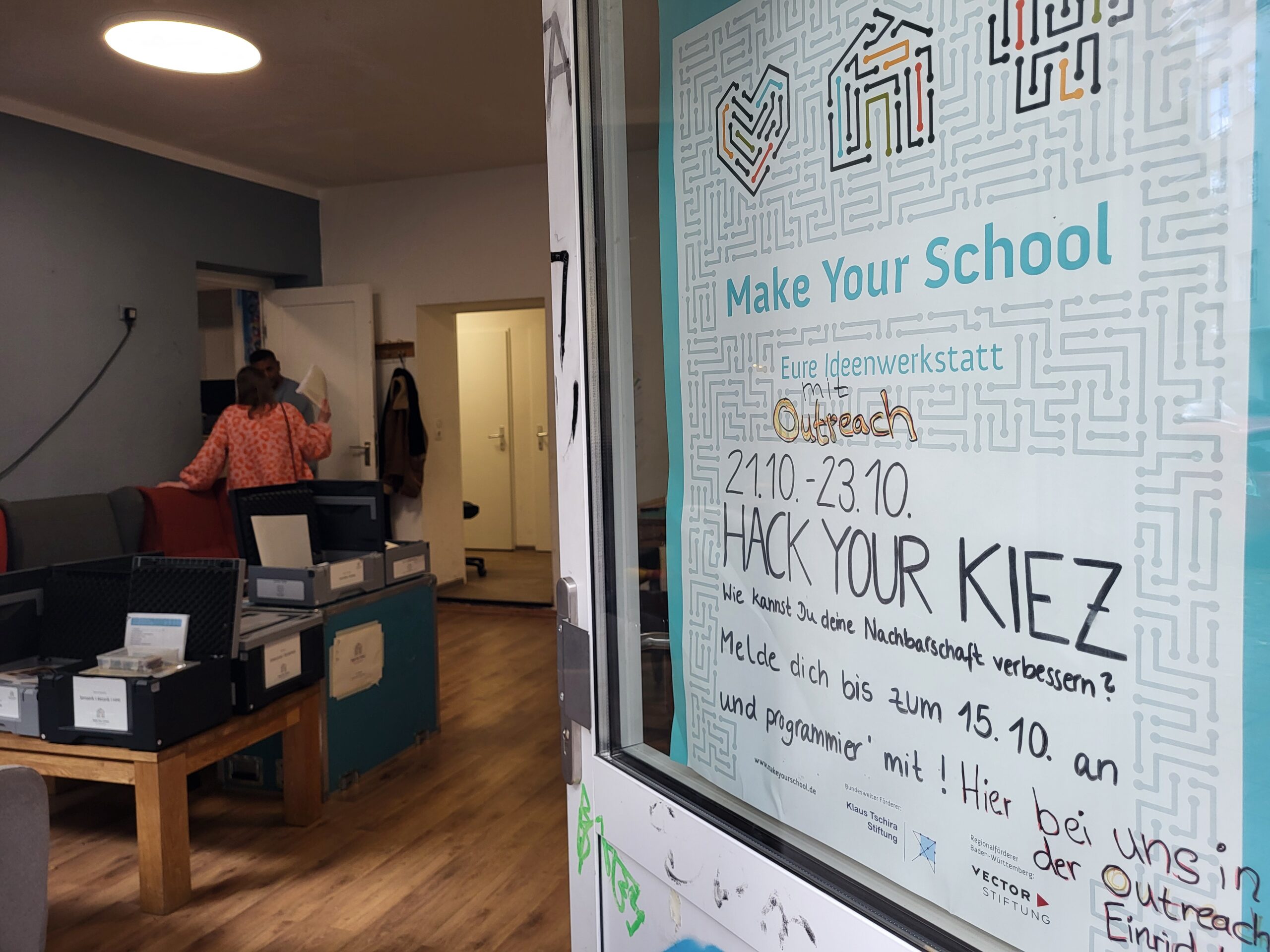 Ein Plakat von Make Your School hängt an der Eingangstür eines Jugendzentrums. Die Tür steht offen und man sieht zwei Menschen im Hintergrund.