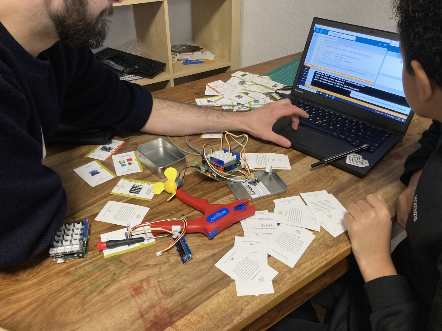 Ein Erwachsener und ein Jugendlicher coden an einem Laptop. Vor ihnen auf dem Tisch liegen Materialkarten und der Prototyp von einem Hack.