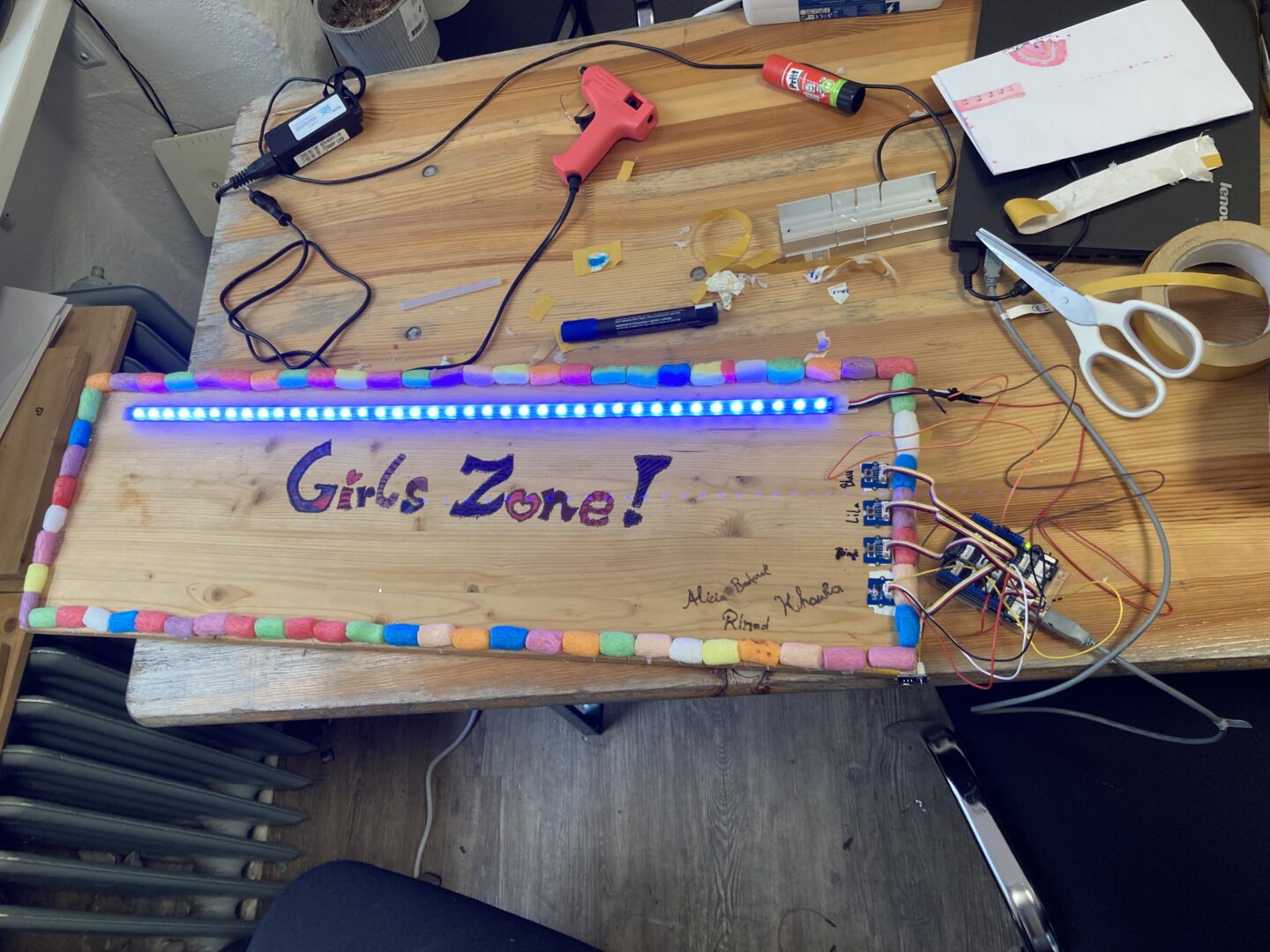 Ein mit LED-Streichen beschmücktes Schild, auf dem "Girls Zone!" steht. Es ist über Kabel an einen Laptop angeschlossen.