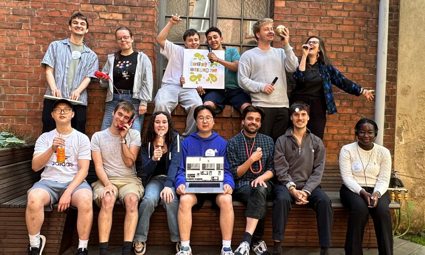 Eine Gruppe von 14 Personen posiert mit Musikinstrumenten vor einer Backsteinwand