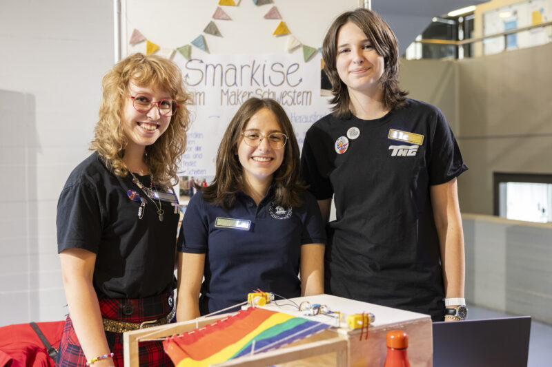 Drei weibliche Jugendliche stehen und lächeln in die Kamera. Hinter ihnen ist eine Stellwand, auf der steht "Smarkise". Vor ihnen ist ein Prototyp: ein großes Gehäuse aus Holz mit einer regenbogenfarbenen Markise.