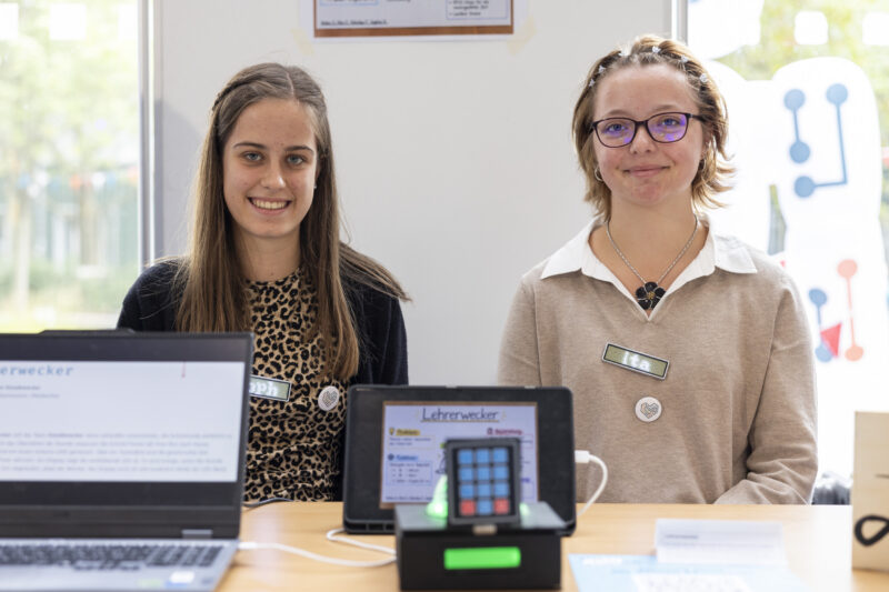 Teamfoto: Zwei weibliche Jugendliche sitzen und lächeln in die Kamera. Vor ihnen ist ein Tablet mit einem Bild, auf dem Bild steht "Lehrewecker", vor dem Tablet ist ein Prototyp zu sehen.