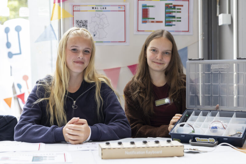 Teamfoto: Zwei weibliche Jugendliche sitzen an einem Tisch und lächeln in die Kamera. Ein Mädchen hält eine offene Box. Vor ihnen auf dem Tisch steht ein Prototyp mit dem Namen "Notification Keyboard".