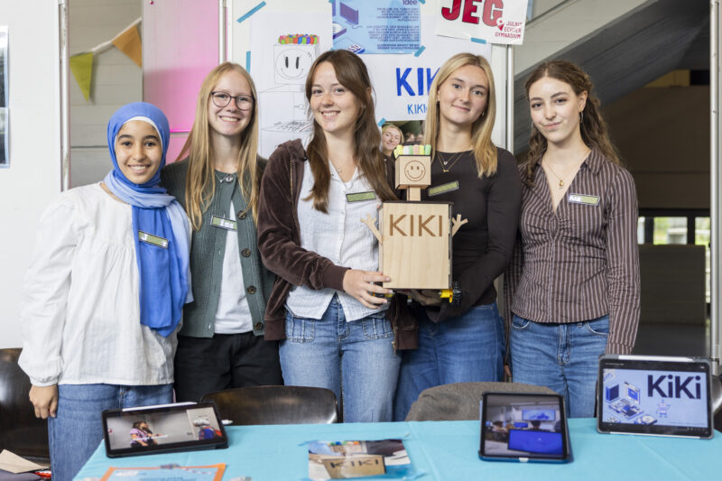 Foto vom Team KIKI: 5 weibliche Jugendliche stehen und lächeln in die Kamera, zwei Mädchen halten einen Roboter aus Holz, auf dem steht KIKI.