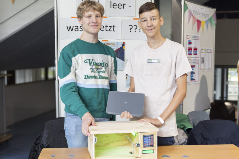 Teamfoto: Zwei männliche Jugendliche stehen und lächeln in die Kamera. Einer hält ein Tablet, der andere legt seine auf auf ein Gehäuse aus Holz mit einem Tastenfeld.