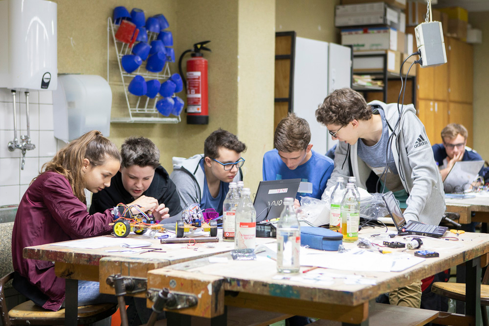 Jugendliche sitzen um einen großen Werktisch herum. Sie stecken die Köpfe zusammen. Zwei basteln an einem Roboter, drei Andere diskutieren vor einem Laptop.